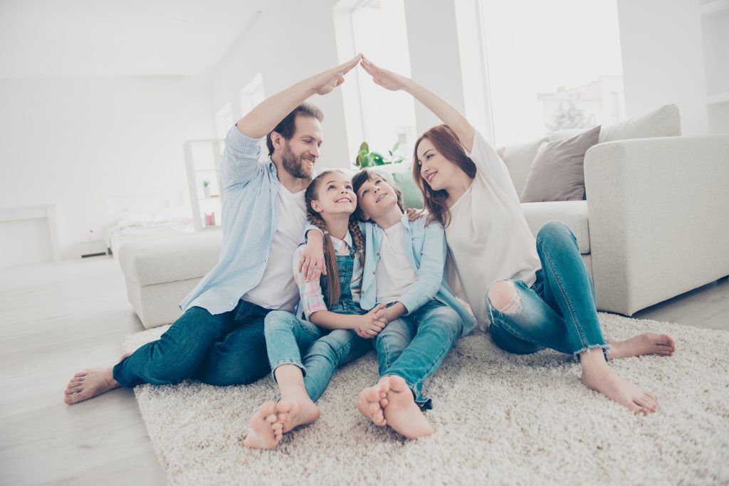 New building residential house purchase apartment concept. Stylish full family with two kids sitting on carpet, mom and dad making roof figure with hands arms over heads